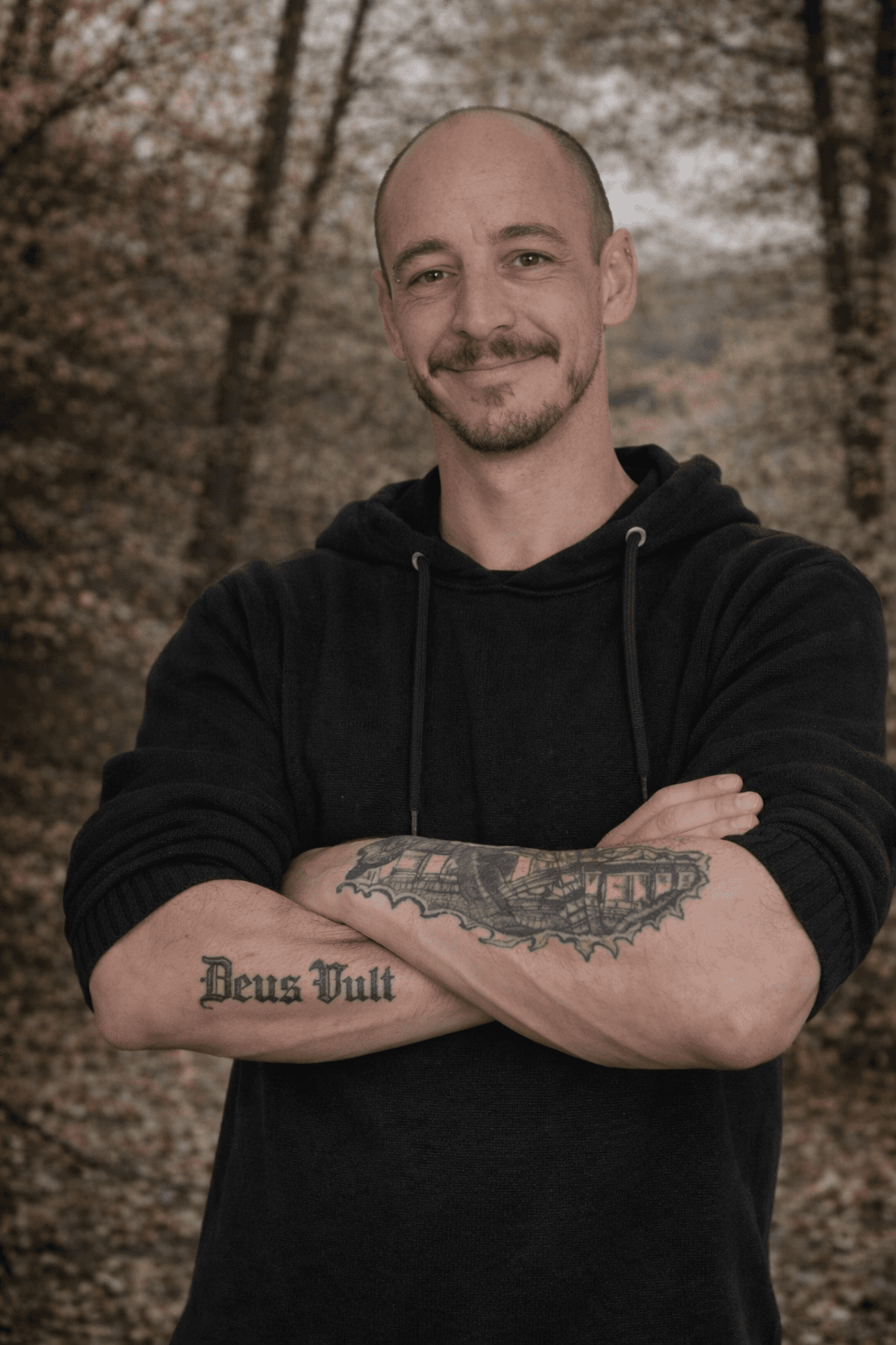 Lächelnder kahler Mann mit verschränkten Armen und Tätowierungen vor einem herbstlichen, verschwommenen Waldhintergrund.